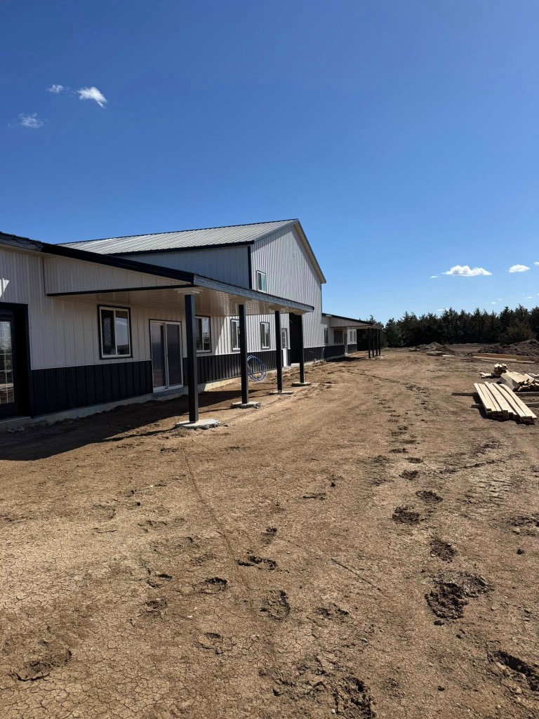 Living wing with deep porch on black posts, glass doors and windows, and taller shop volume beyond under blue sky.