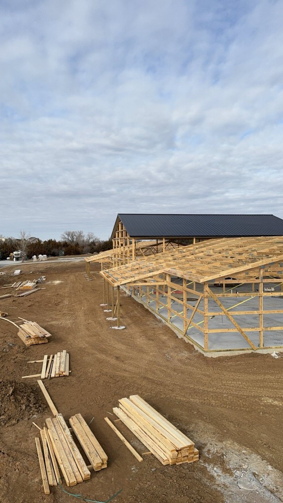 Framing stage: black metal roof on the tall bay, exposed trusses and girts on lower wings, temporary bracing.