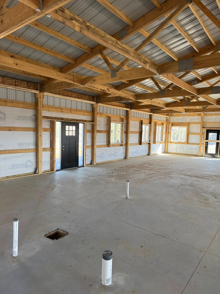 Living side interior: concrete slab with plumbing rough-ins, Barricade wrap, trusses, windows, and entry doors.