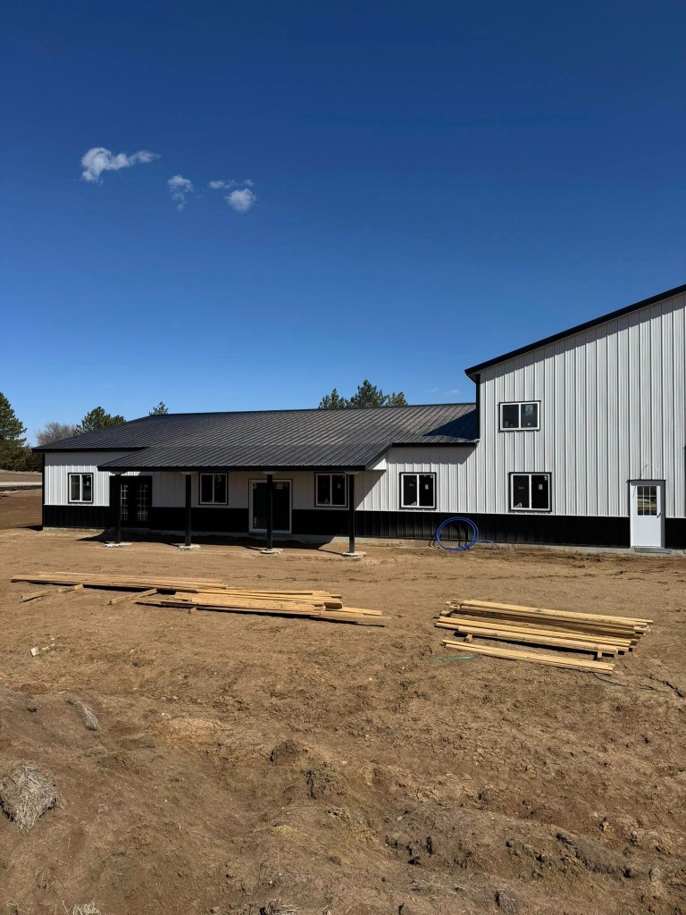 Angled view of the L-shaped shell: porch, stacked lumber on site, and mixed roof heights.