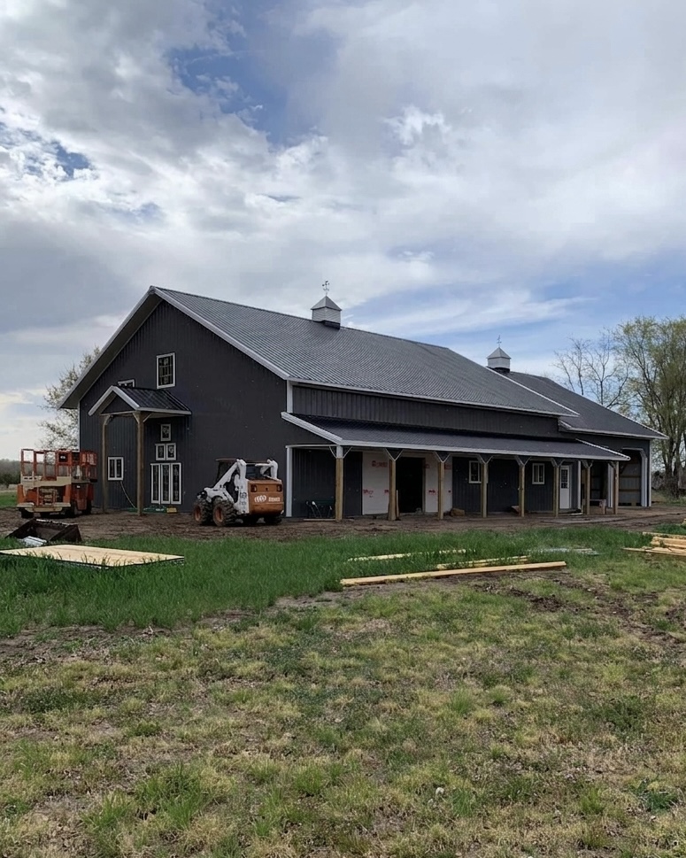 Charcoal metal barndominium post-frame shell with white trim, ridge cupolas, wood porch posts, and equipment on a rural build site.