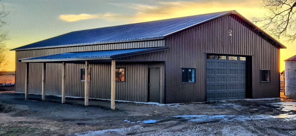 Wide view of a bronze post-frame building with lean-to porch at sunset—cool sky at left, warm golden horizon—Nebraska.