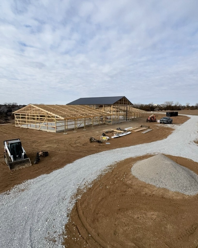 Construction progress of a large multi-wing shouse—slab, post-frame framing, black metal roof on the main bay, and equipment on site.