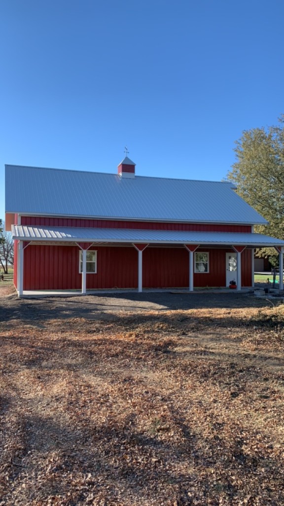 Red vertical metal barn with full-length white-roof porch, white posts with red braces, cupola, and clear blue sky.