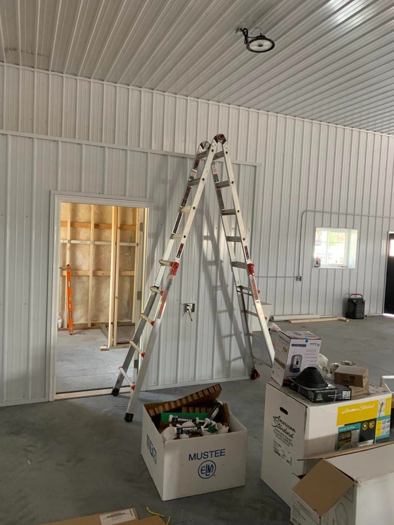 Interior build-out in progress: white metal liner, doorway to insulated stud room, conduit, and plumbing and finish supplies on the slab.