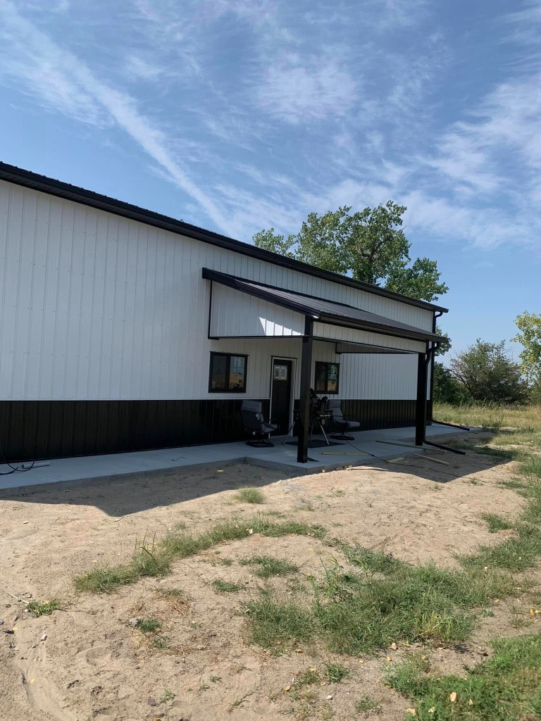 White upper walls and black wainscot post-frame shop with lean-to porch, black posts, and sliding windows under cover.